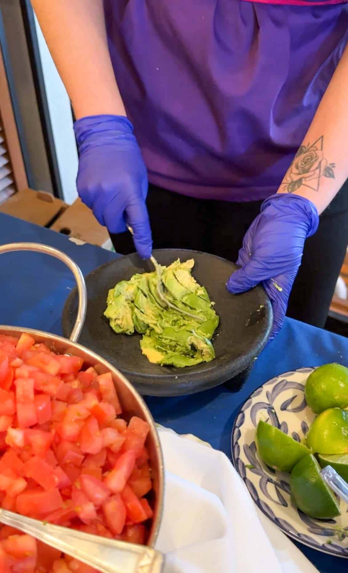 Hands with purple plastic gloves mashing avocado in a black bowl with a fork on a blue tablecloth.