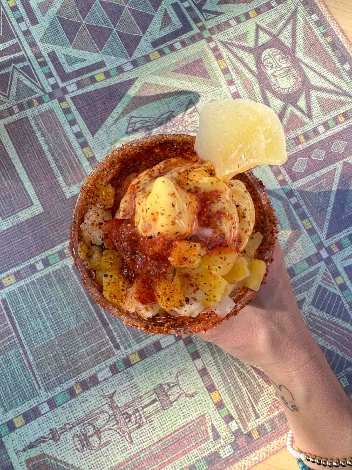 Top view of a hand holding a chile mango flavor dole whip with a candied mango on the rim on a mauve decorated table.