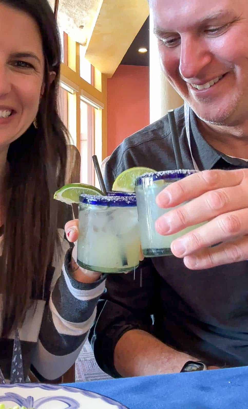 A man and woman holding margaritas in clear blue rimmed glasses and doing cheers with them.