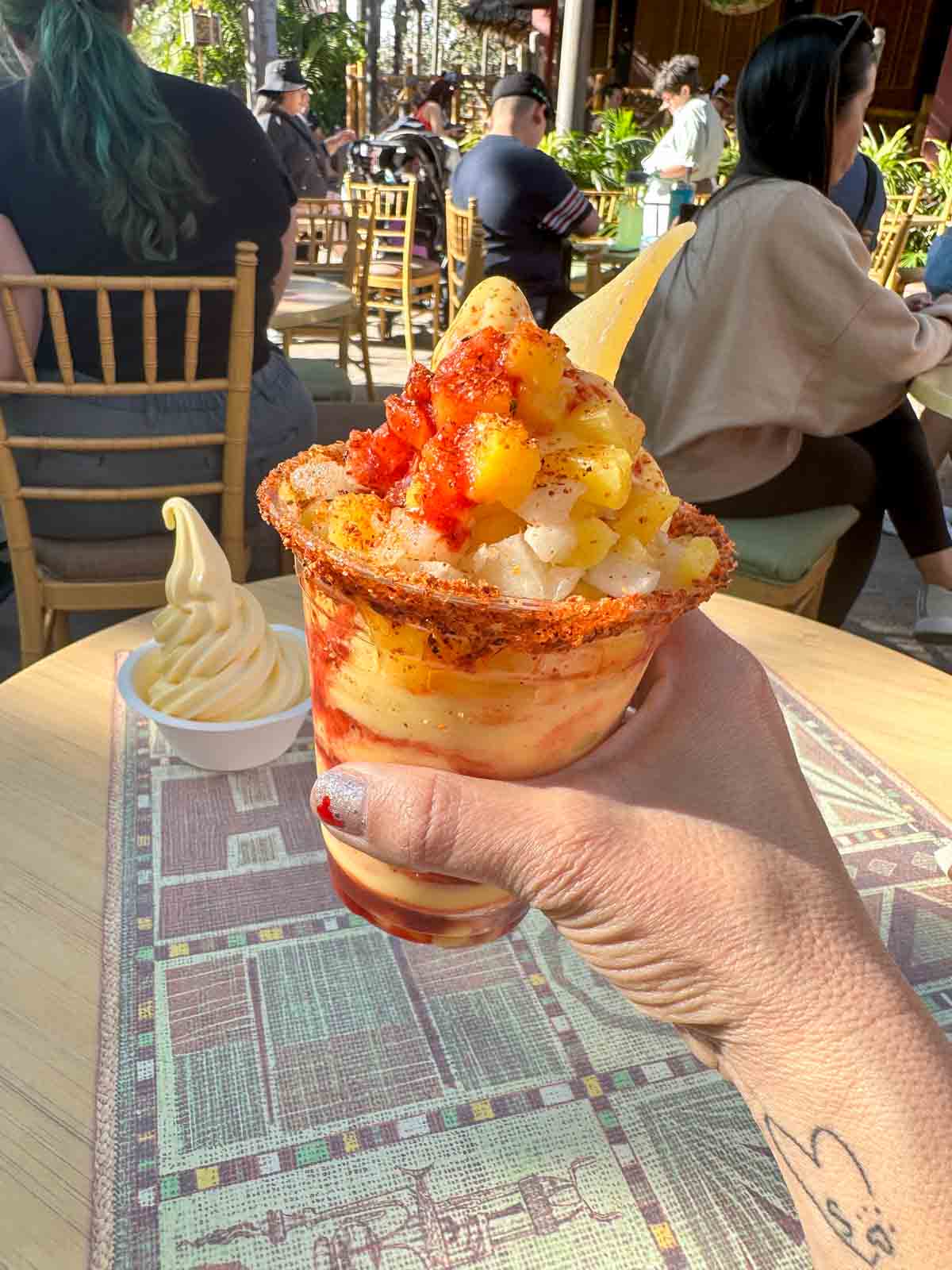 A hand holding a mango chile flavored dole whip over a table at Disneyland.