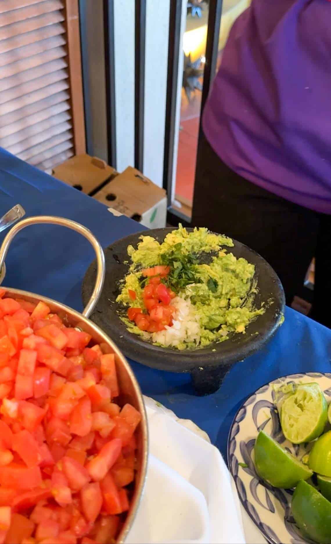 Mashed avocado topped with chopped onions, tomato and cilantro in a black bowl on a blue tablecloth.