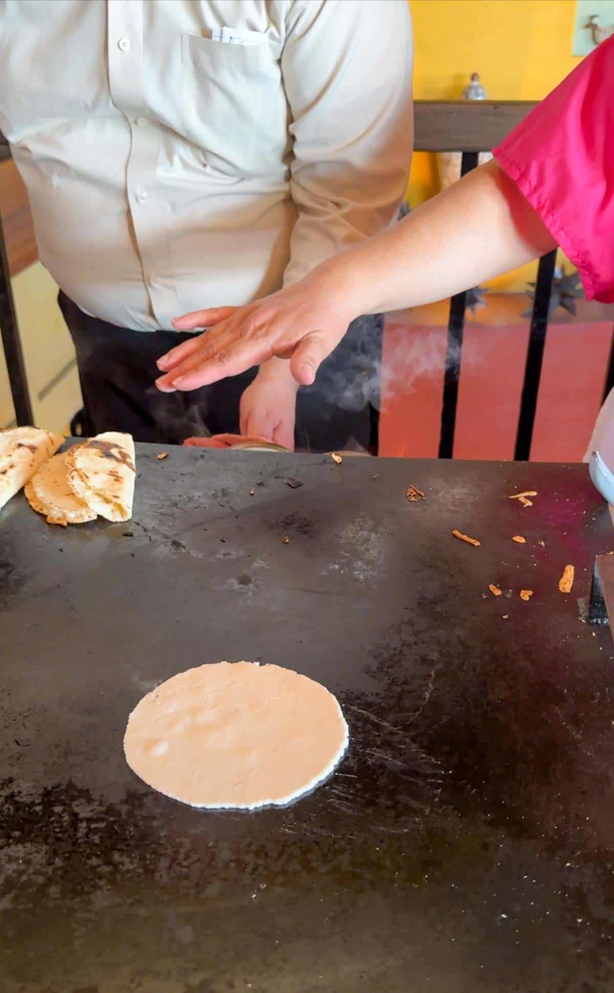 A raw corn tortilla on a large flat grill top with a womans hand over it.