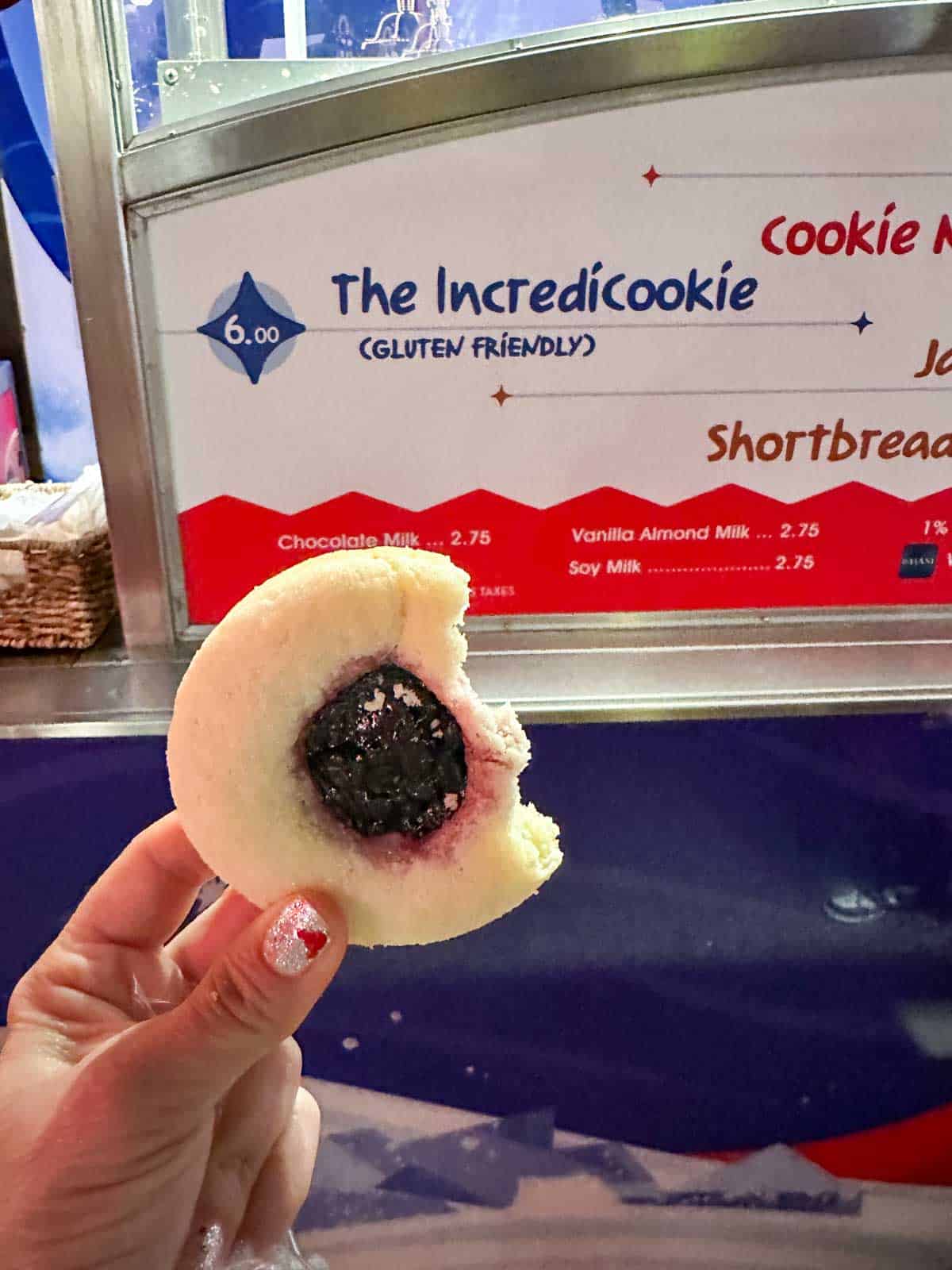 A hand holding a sugar cookie with a purple fruit middle in front of the cookie sign that has a bite out of it.