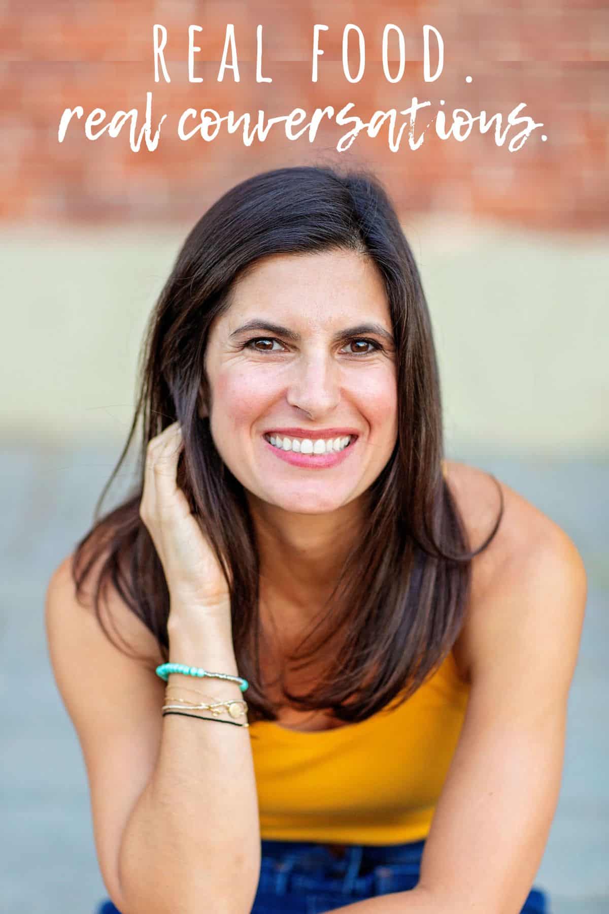 A title of a podcast called Real Food Real Conversations in white over a brick wall and a woman in yellow below it holding back her hair and smiling.