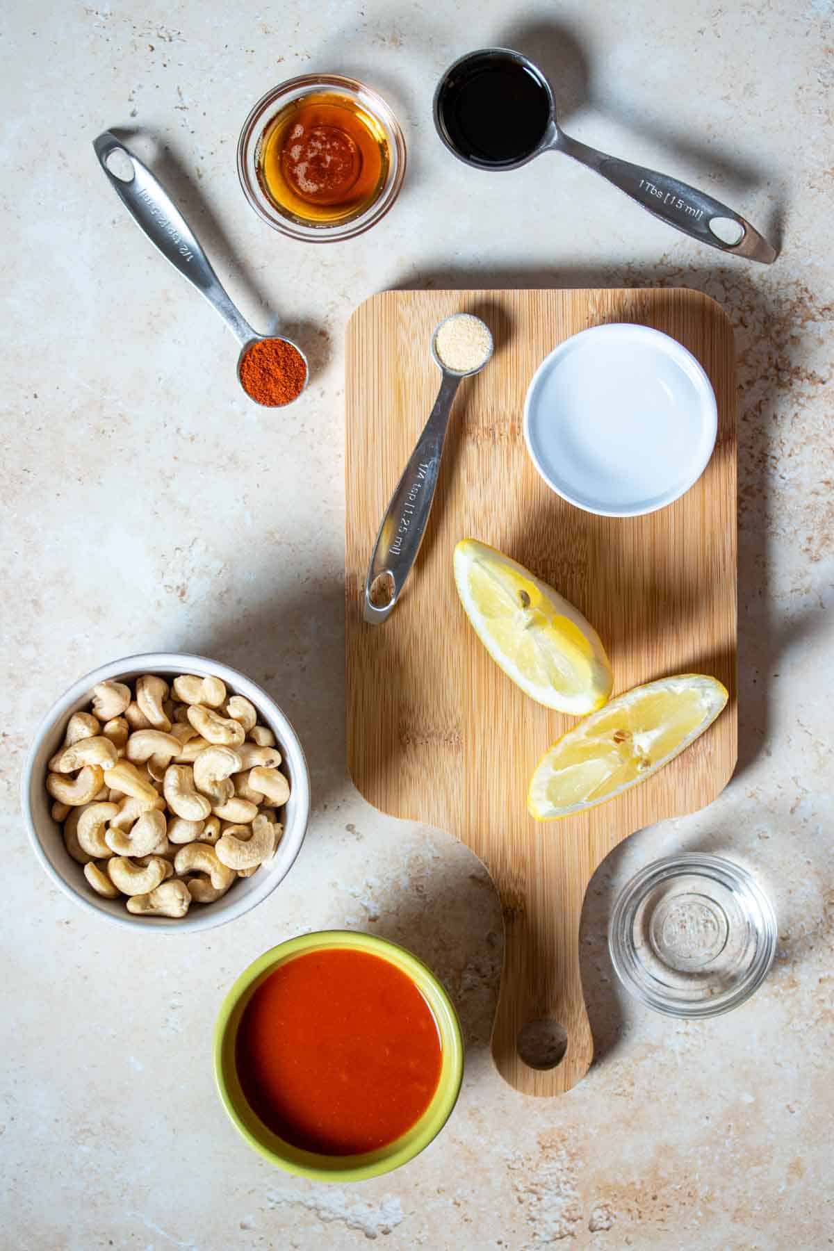Top view of a tan surface with a cutting board and cashews, a red sauce in a bowl, lemon and other spices and liquids in small bowls and spoons.