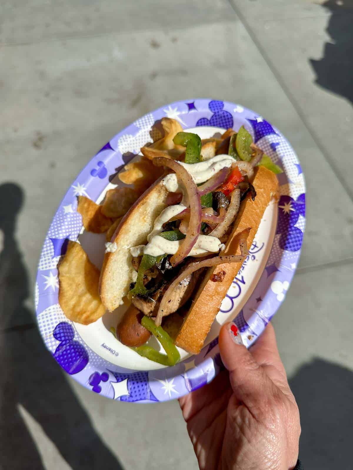 A hand holding a paper plate with a sausage in a bun covered with grilled peppers and white sauce.