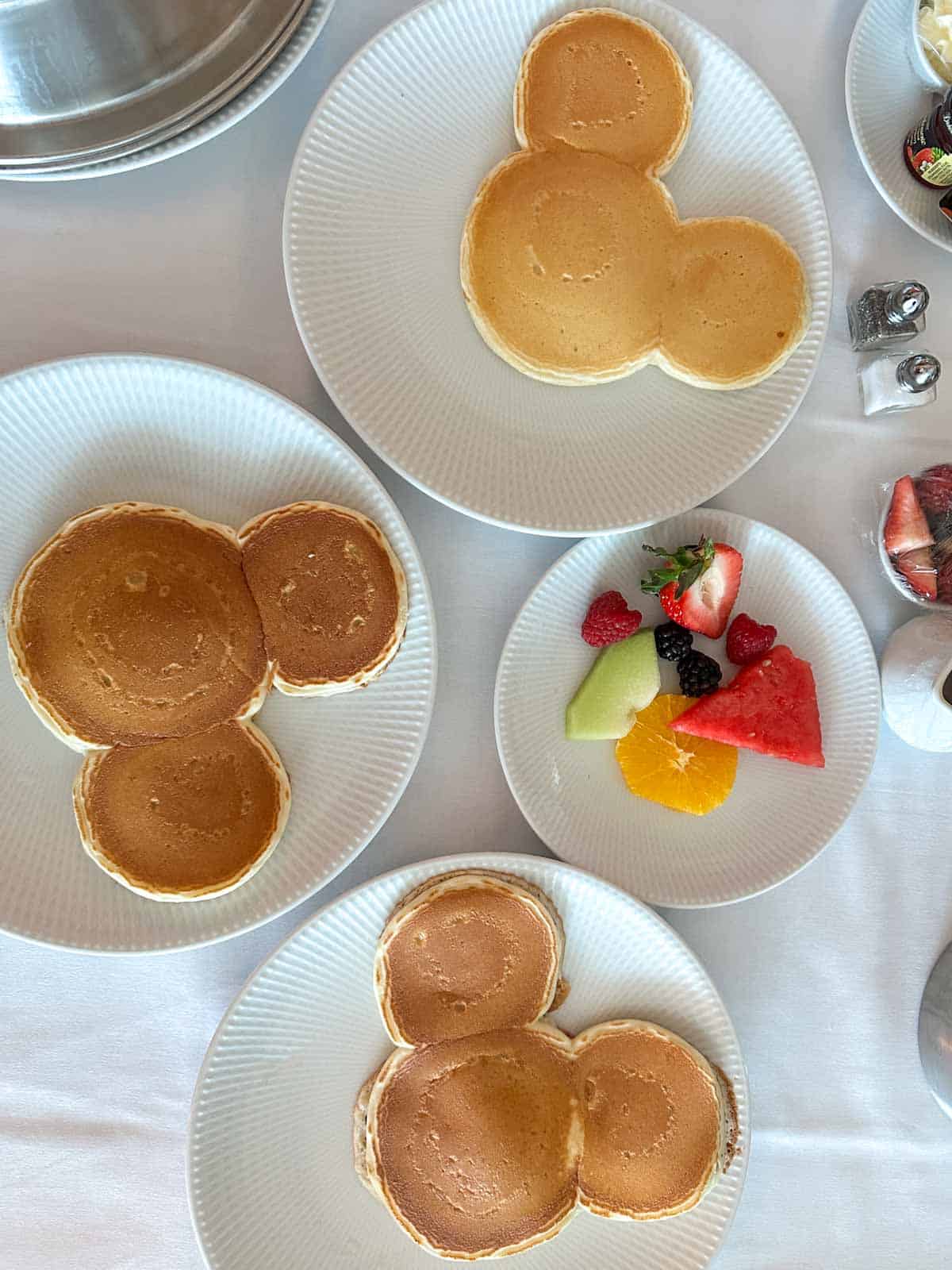 Top view of three white plates with mickey pancakes on them next to a plate of fruit.