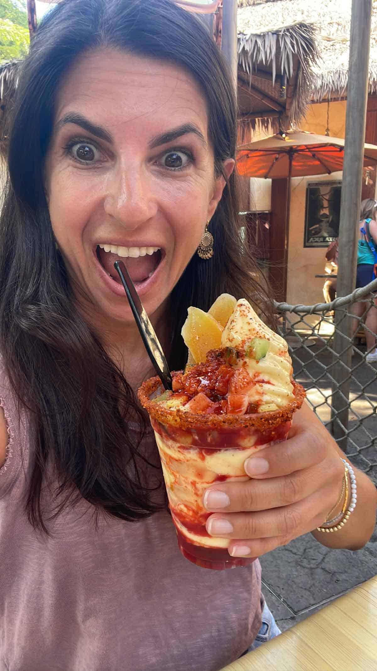 A woman in a pink tank top holding a mango topped pineapple Dole whip with a red sauce on it.