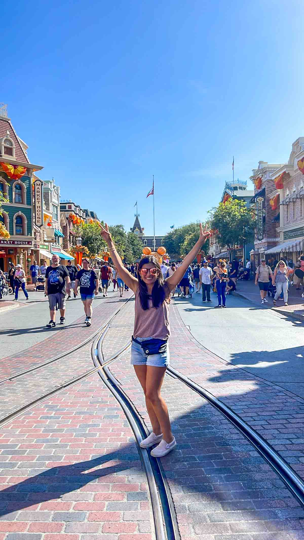 A woman in a pink tank top and cut off jean shorts with her hands up standing in the middle of Main Street Disneyland.