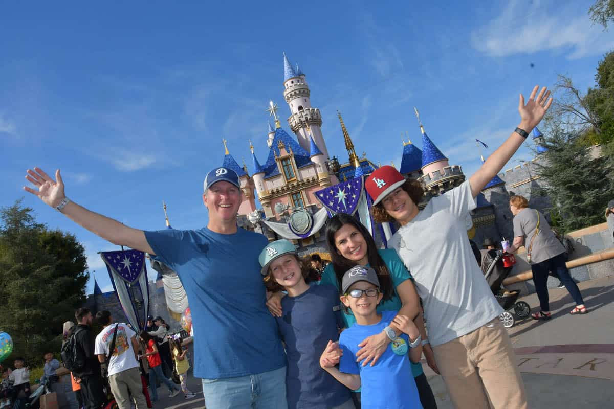 A family of five hugging and the two people on the end holding their arms out in front of the castle at Disneyland.