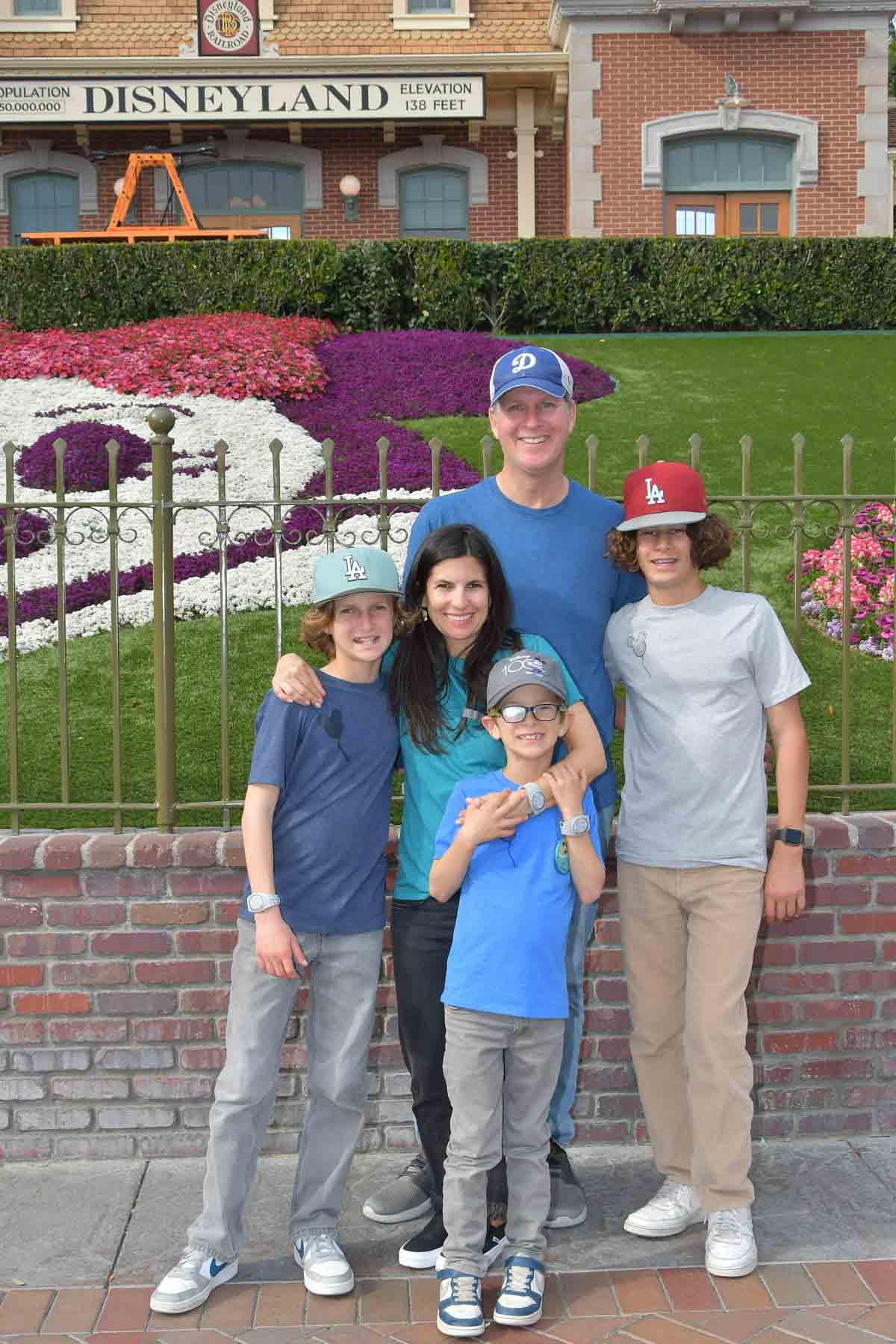 A family of five in blue t shirts with one in grey standing together in front of the flower mickey at the front of Disneyland.