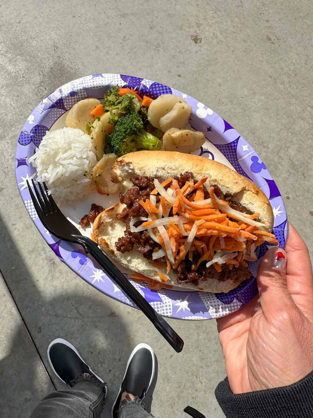 A hand holding a paper plate with a ground meat sandwich in a bun next to rice and vegetables.