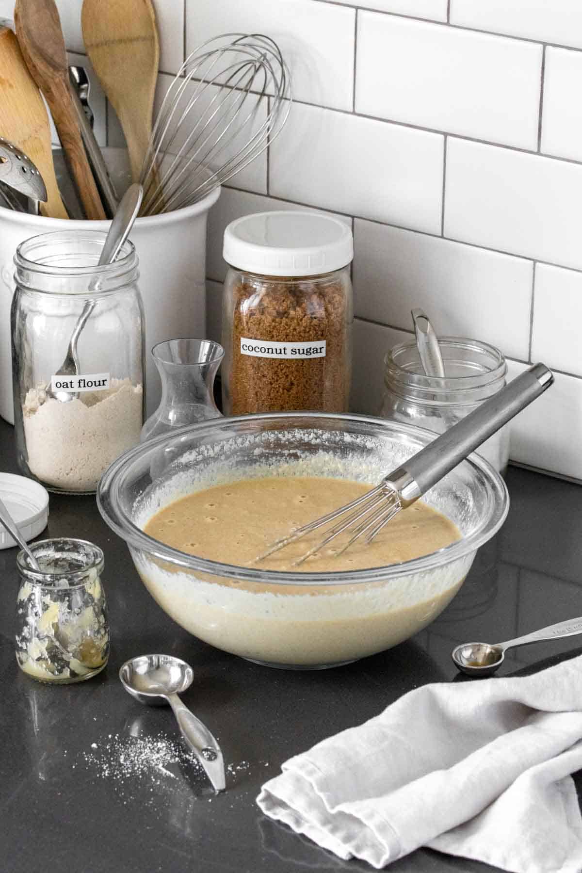 A glass bowl with light tan batter and a whisk in it on a grey kitchen countertop surrounded by jars of sugar.
