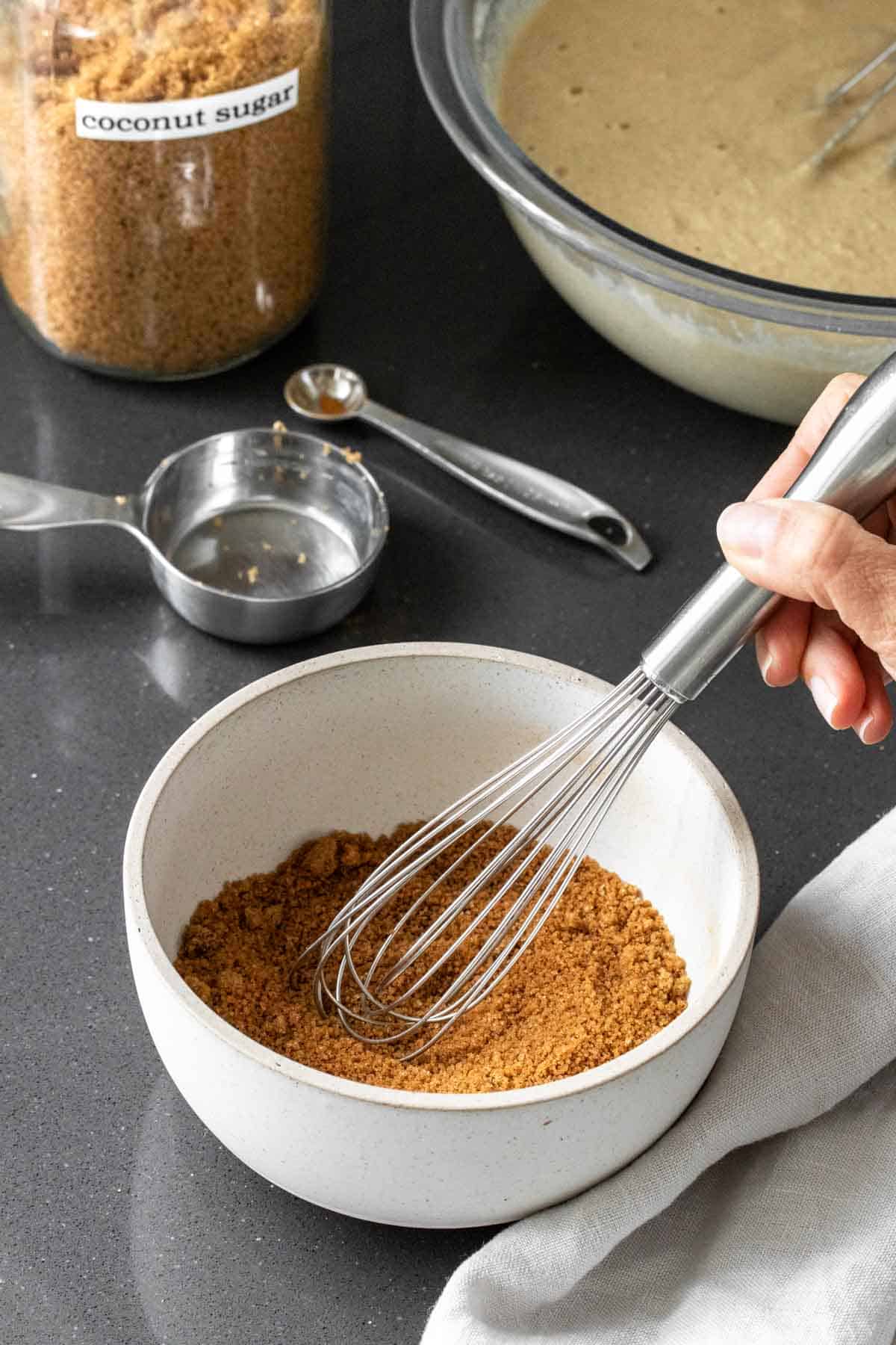A hand with a whisk mixing brown sugar in a white bowl in front of a bowl of cake batter.