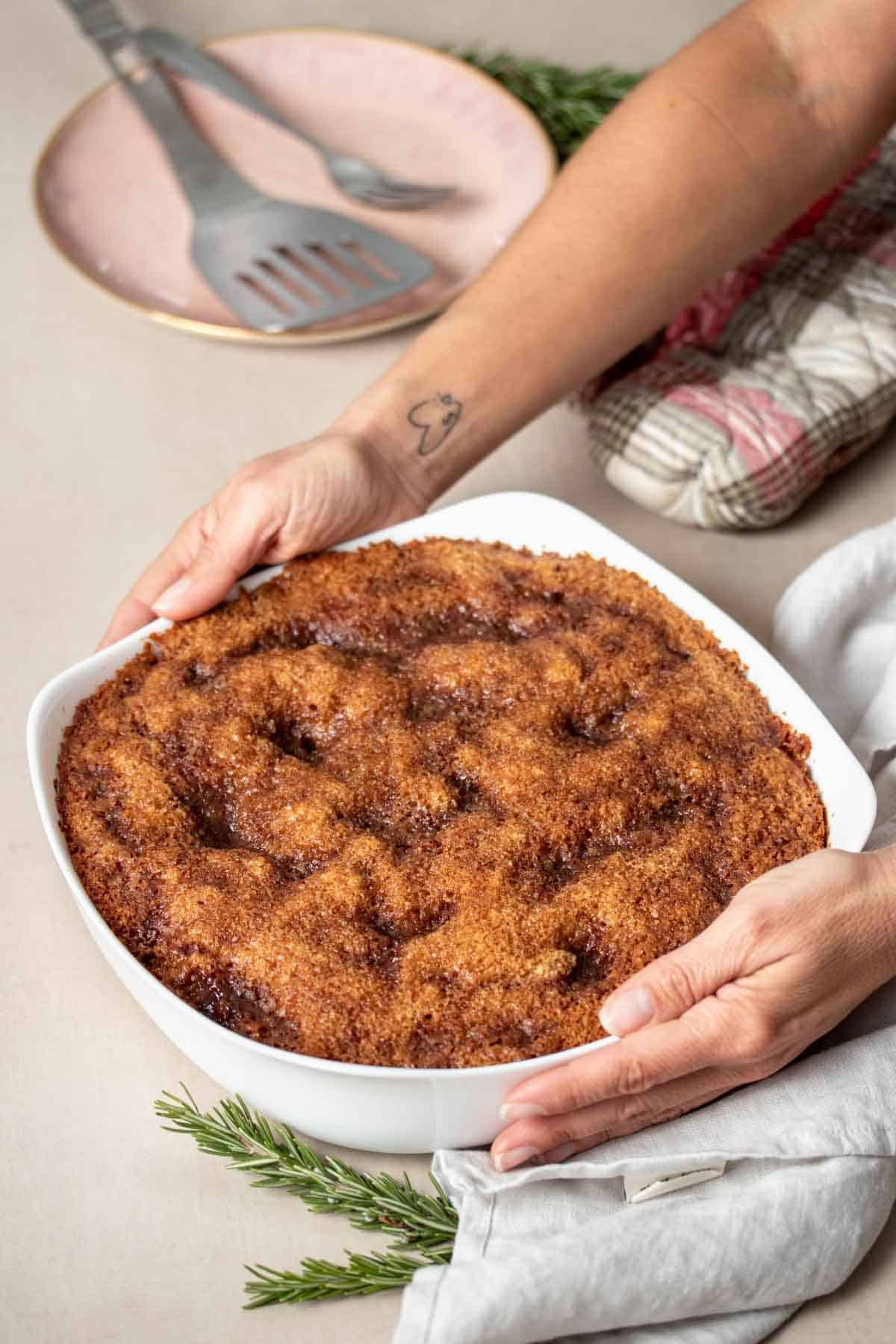 Hands holding a white baking dish with baked coffee cake in it and setting it on a tan surface with a grey towel.