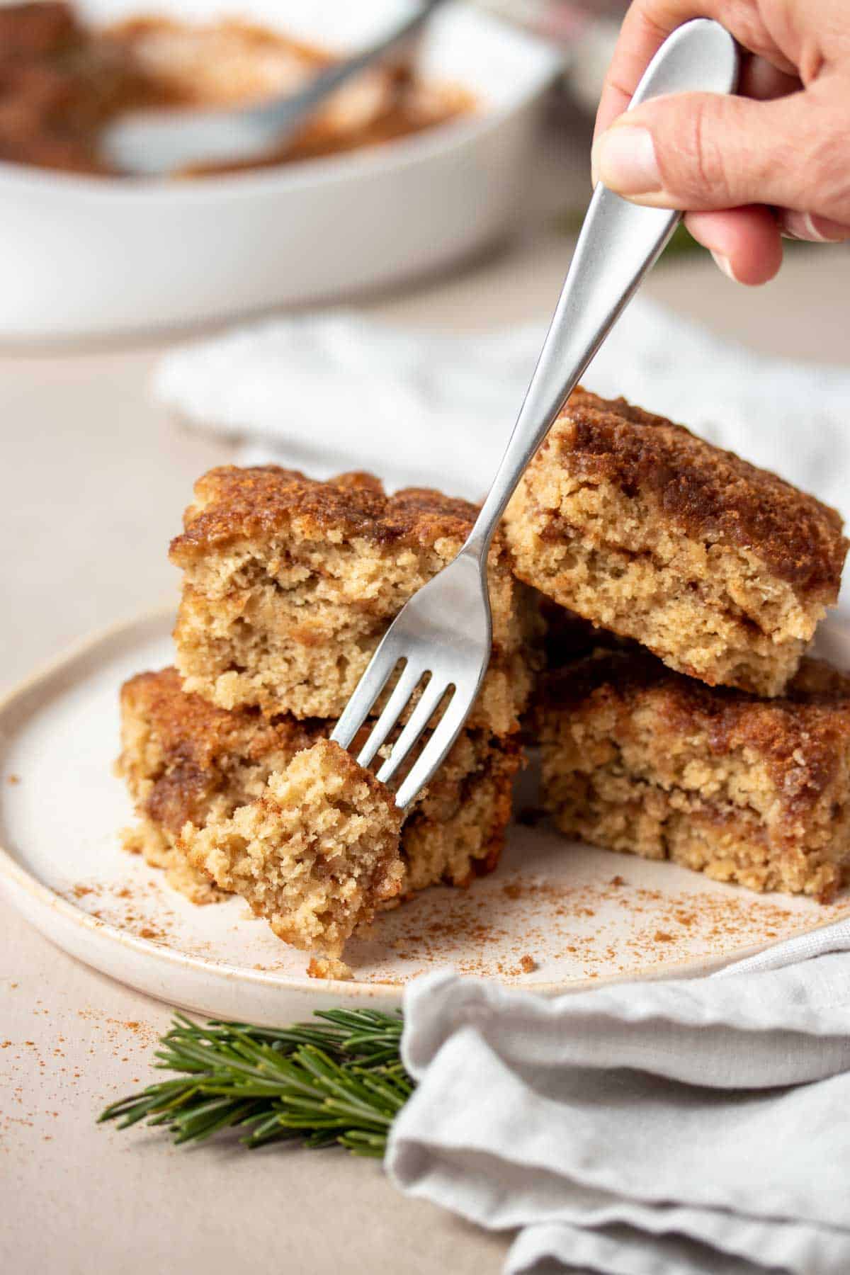 A fork getting a bite from a piece of coffee cake that is piled on a cream plate sitting on a white towel.