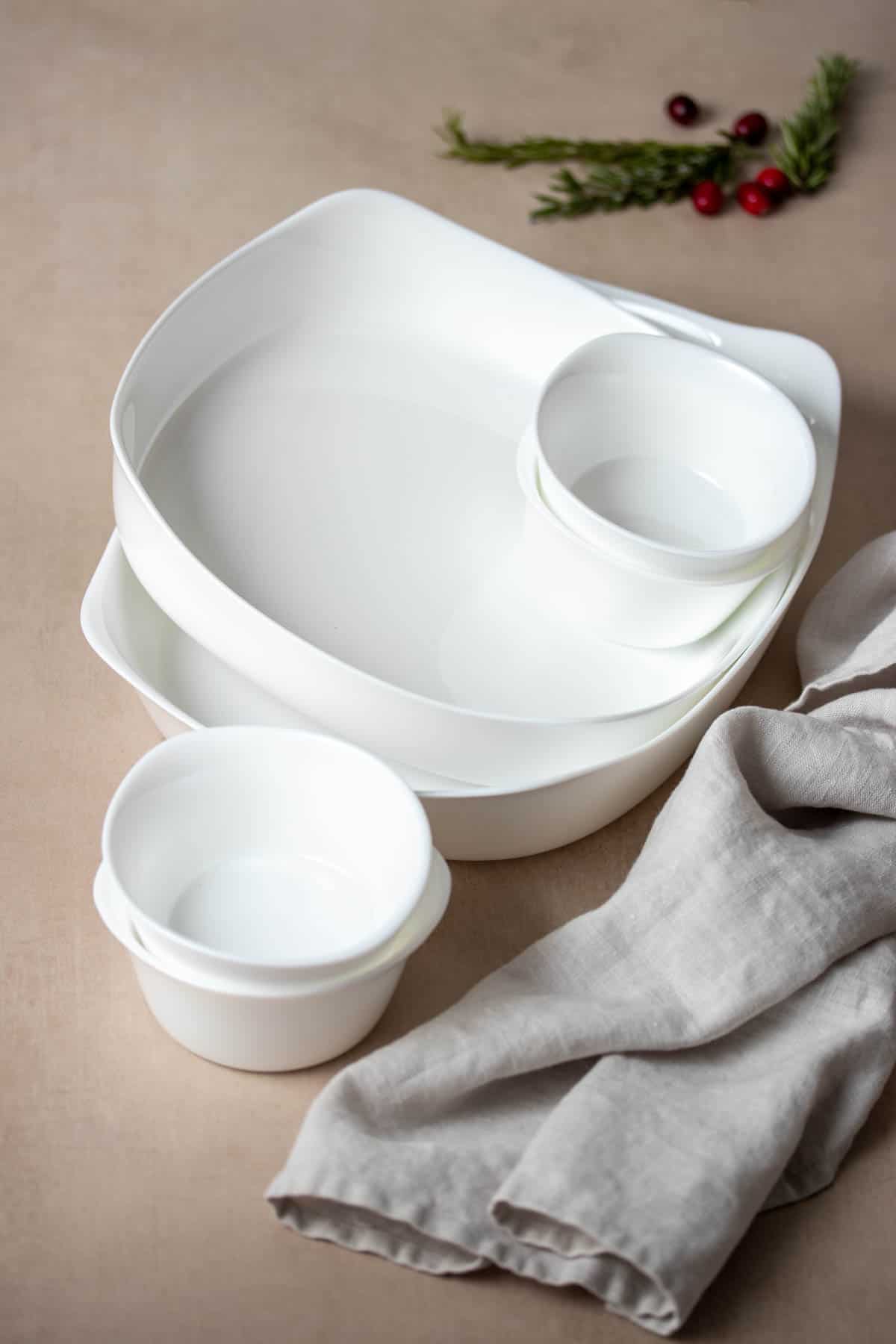 A variety of white baking pans in small to large stacked on a tan colored surface with a grey towel next to them.