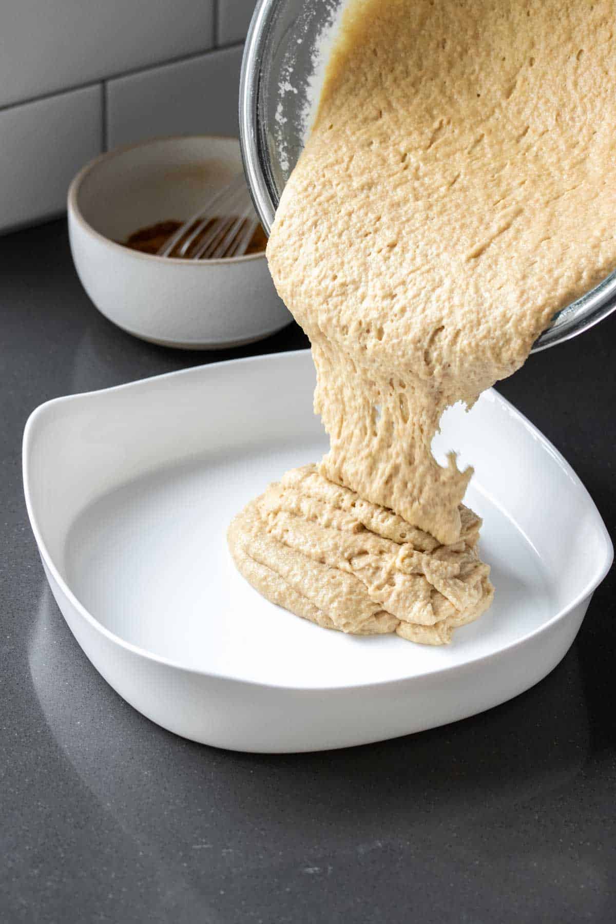 Cake batter being poured from a glass bowl into a white cake pan sitting on a grey kitchen counter.