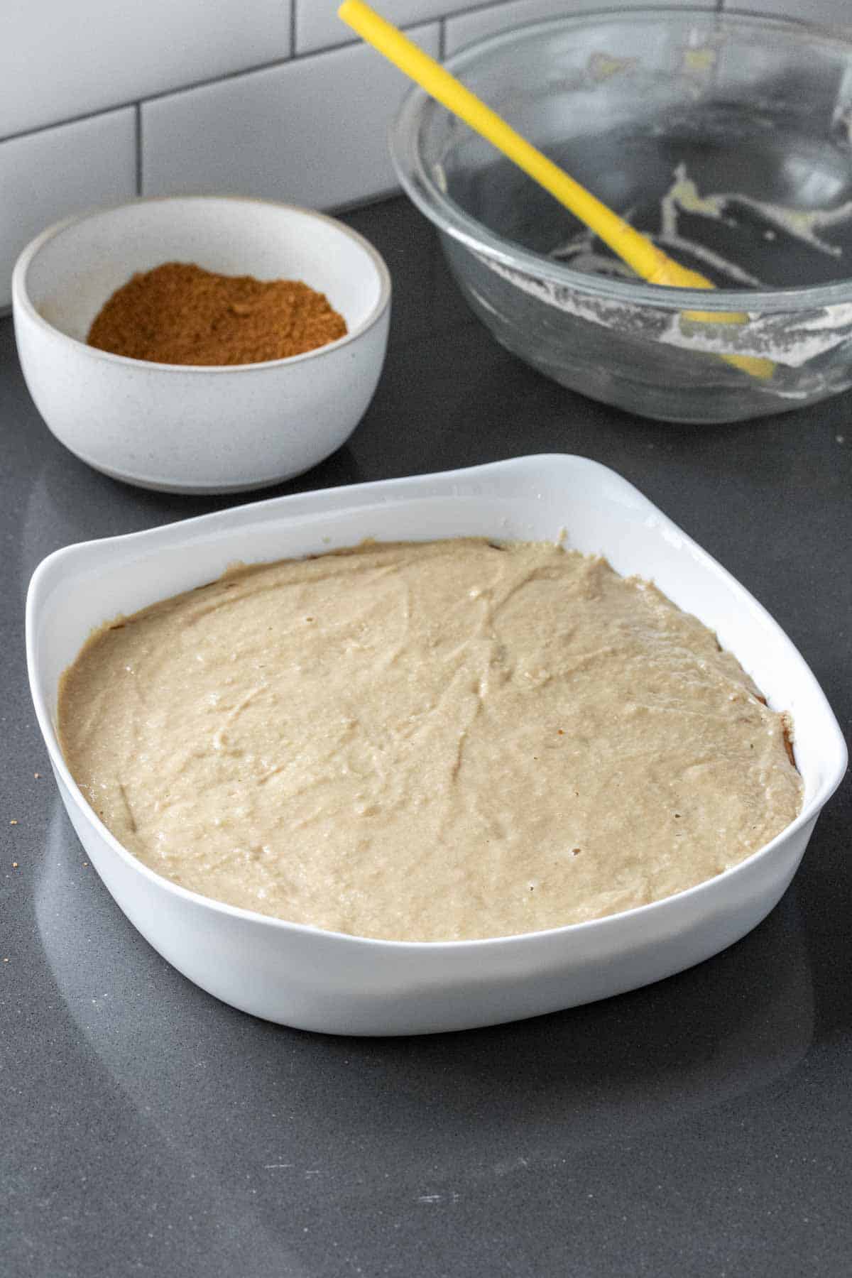 Cake batter in a white cake pan on a kitchen counter in front of a white bowl and an empty glass bowl.