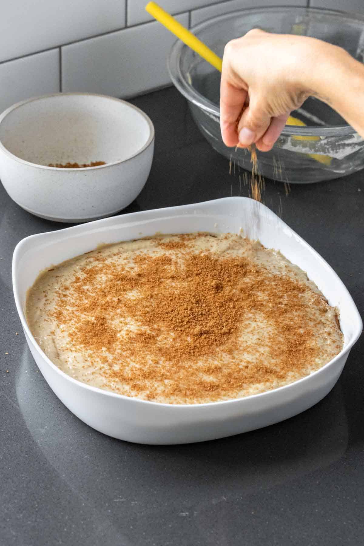 A hand dropping brown sugar on top of a cake batter in a white cake pan that is on a kitchen counter.