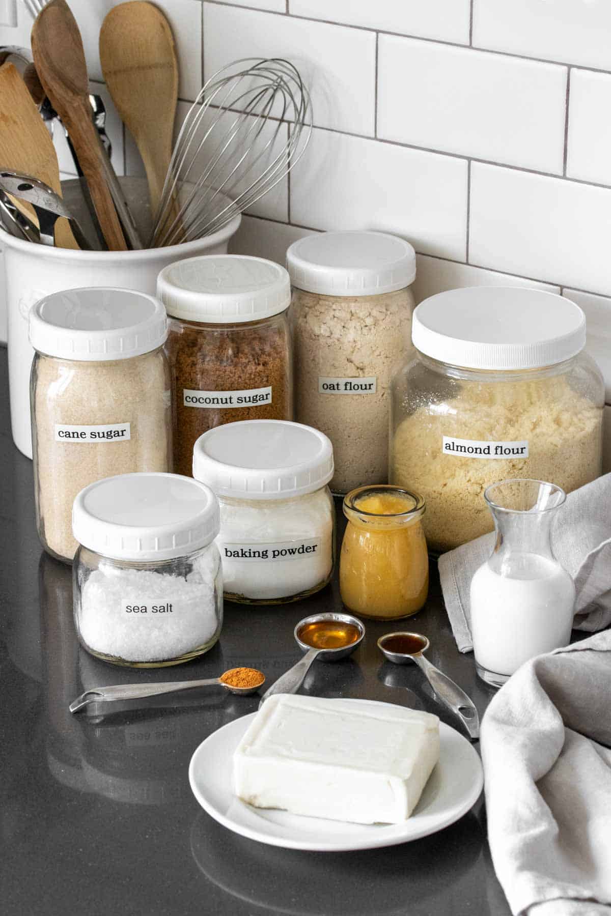 Different sized jars filled with baking supplies and with labels and white tops on a grey kitchen countertop.