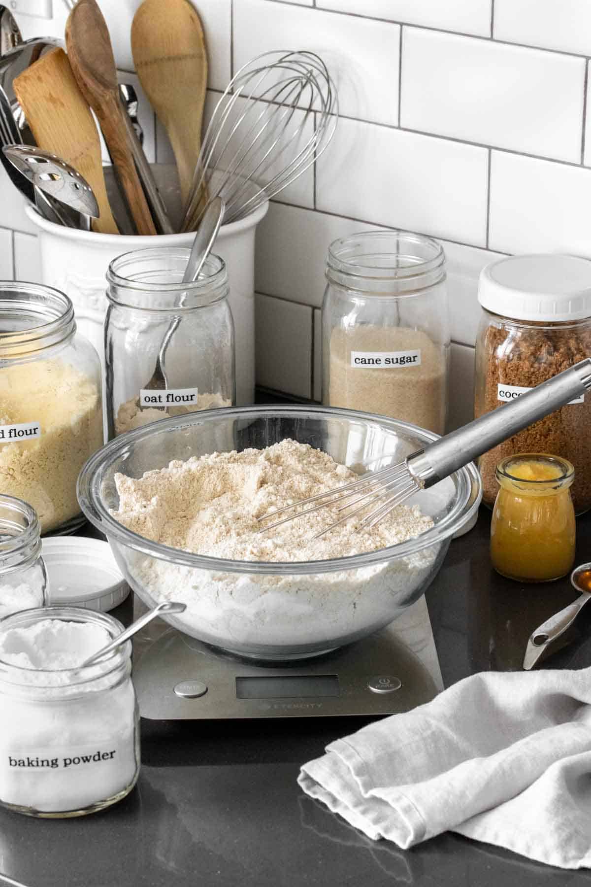 A glass bowl with flour and a whisk sitting on a kitchen scale surrounded by baking supplies on a countertop.
