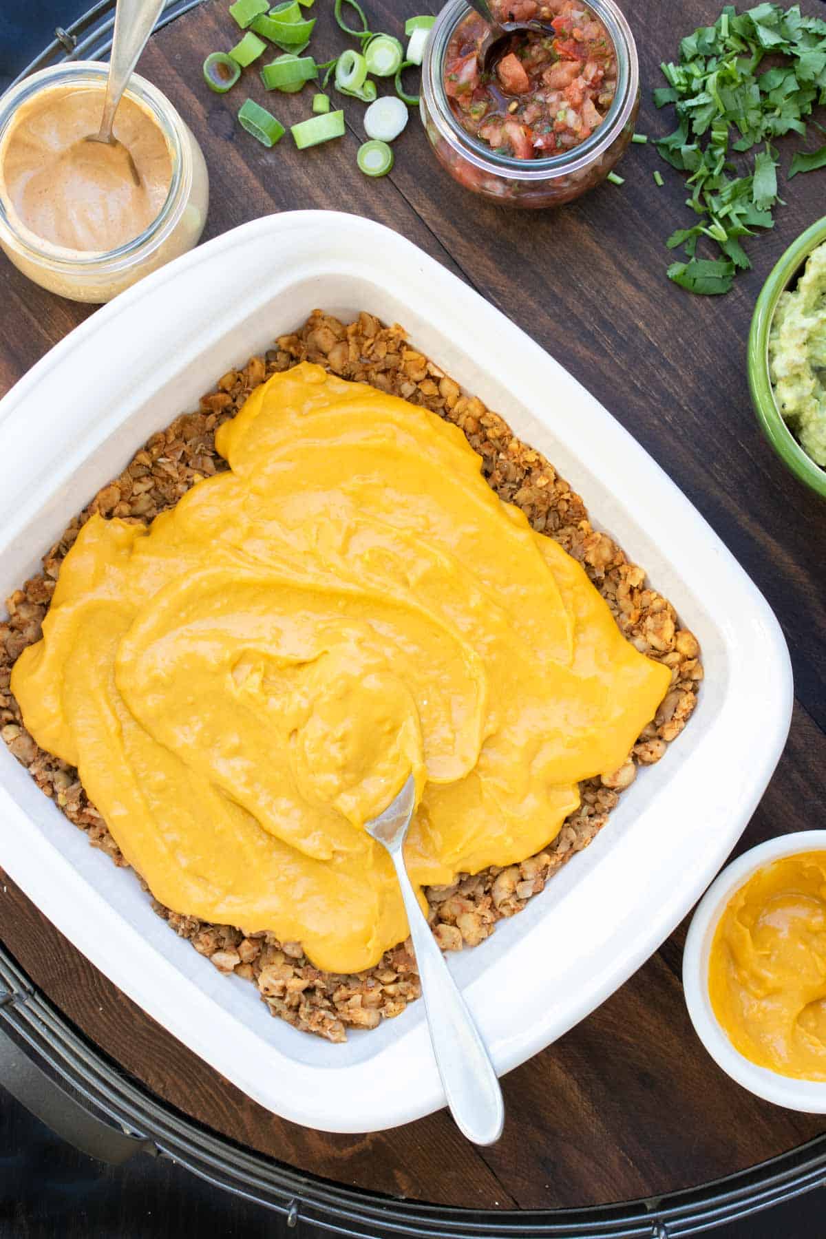 A spoon spread cheese sauce over a layer of taco meat in a white square baking dish.