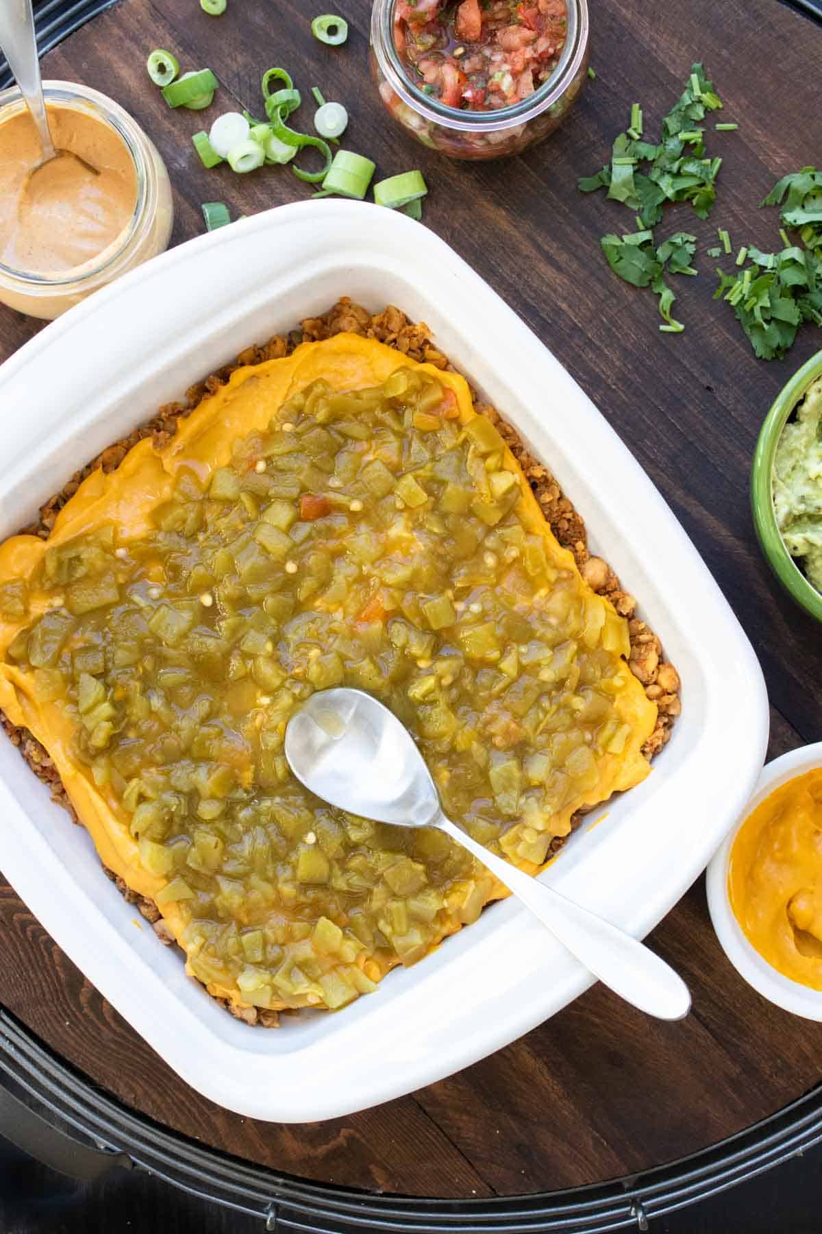 A square white baking dish with taco meat at the bottom mostly covered with cheese sauce and chopped chiles being spread on the top.