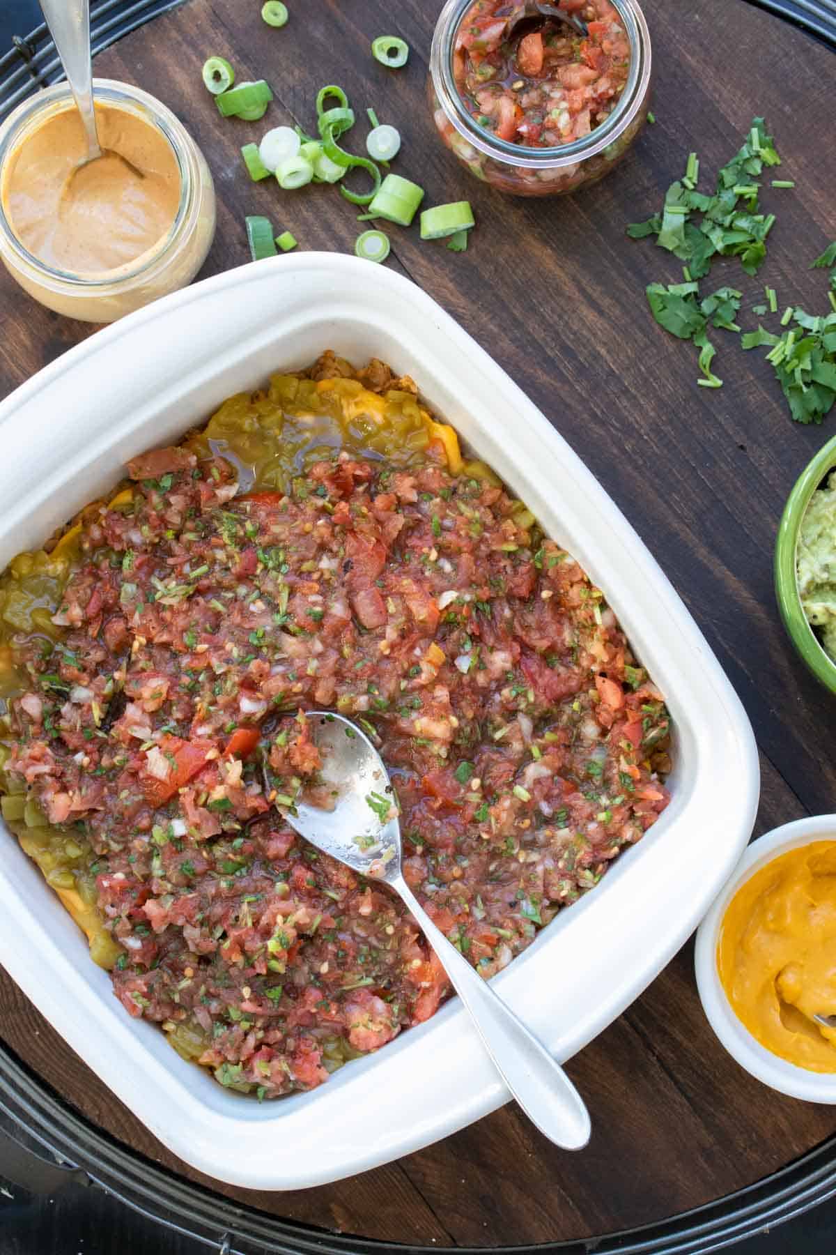 A metal spoon putting salsa over a layer of chiles that is covering more layers of a 7 layer dip.