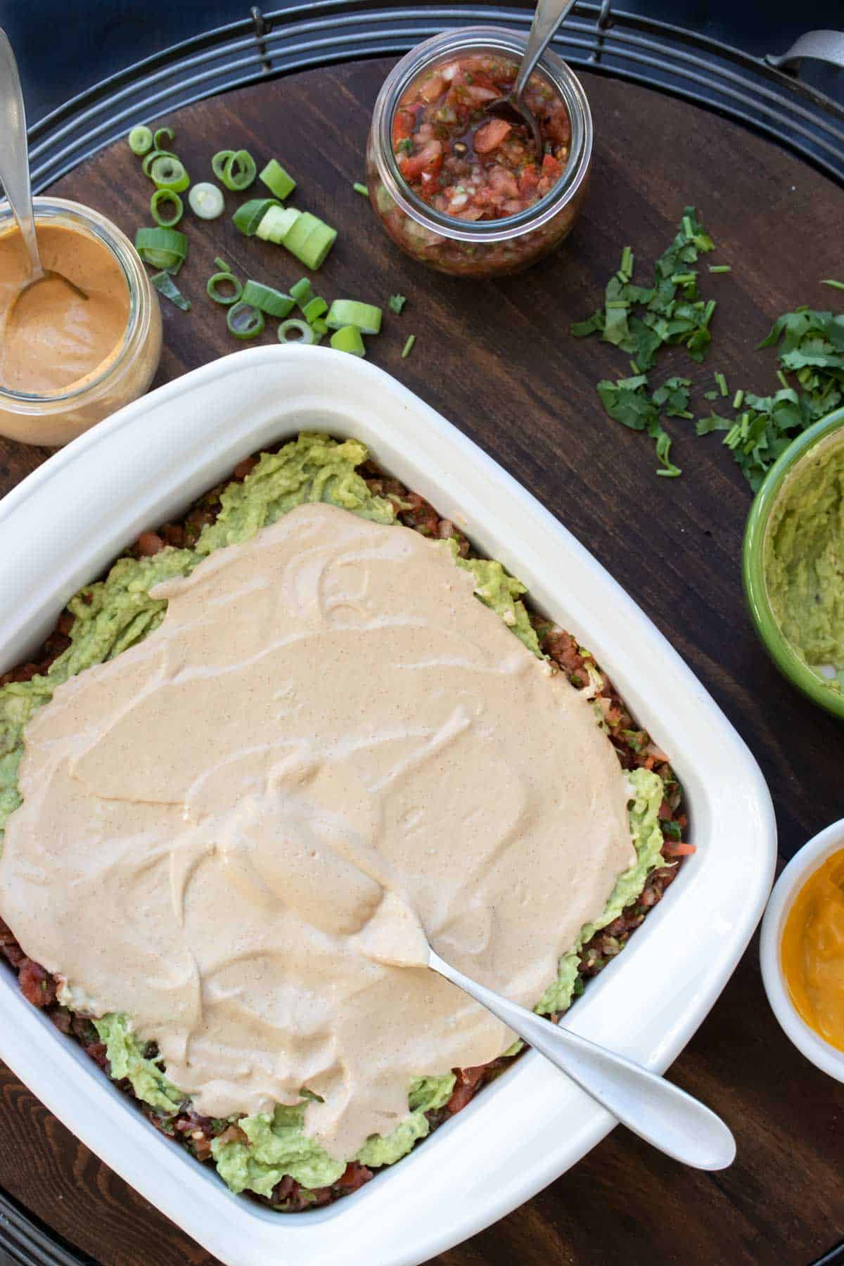 A light pink creamy sauce being spread over a layer of guacamole that is the top of a 7 layer dip in a baking dish.