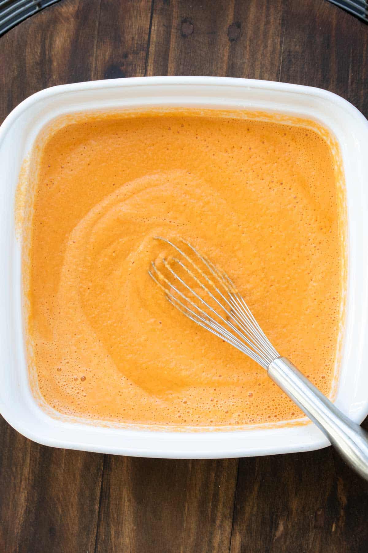 A whisk mixing a buffalo sauce mixture in a square white baking dish.