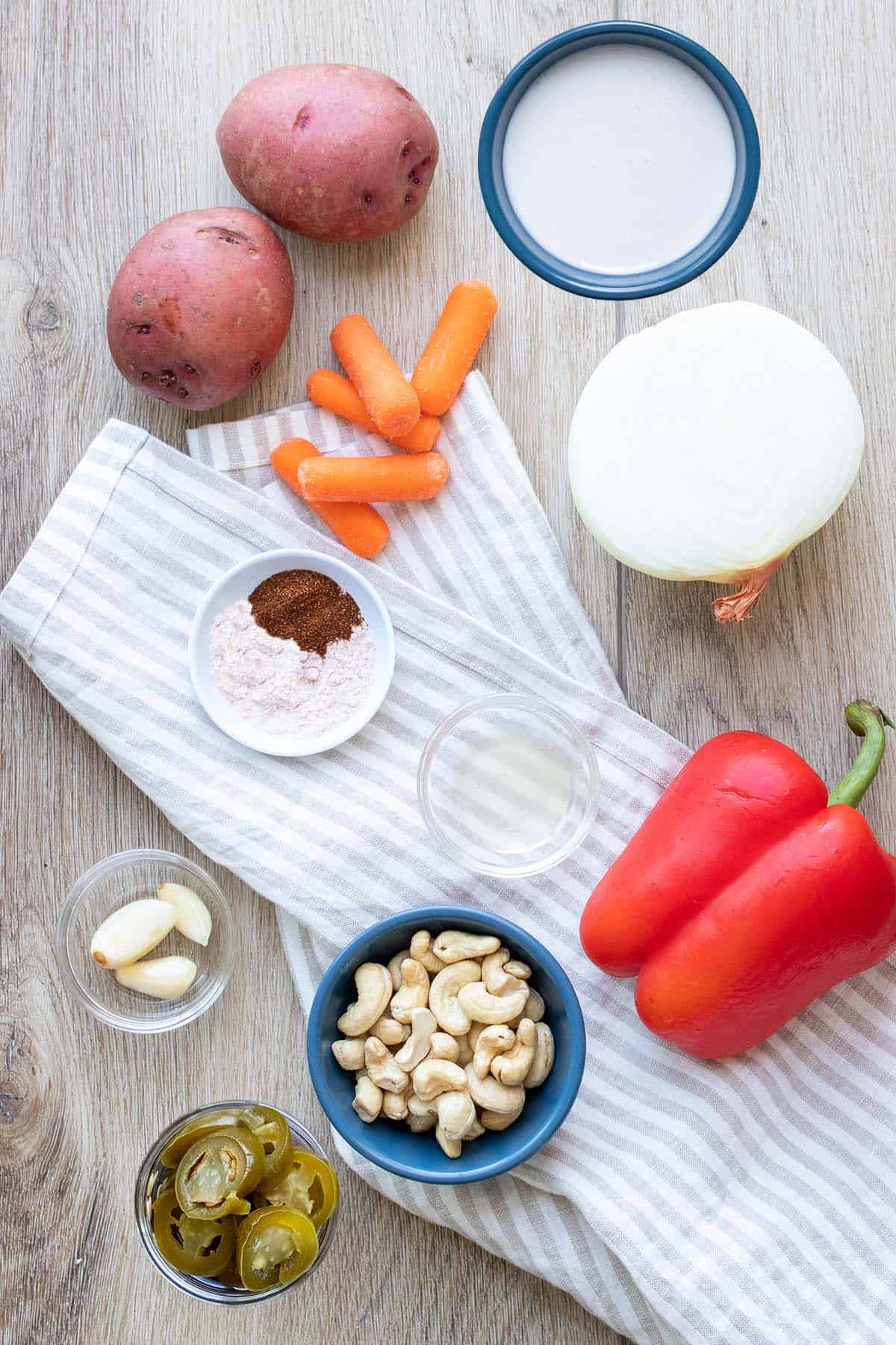 Veggies on a light wooden background next to bowls of spices, garlic, milk, cashews, nacho slices and a clear liquid on a striped towel.