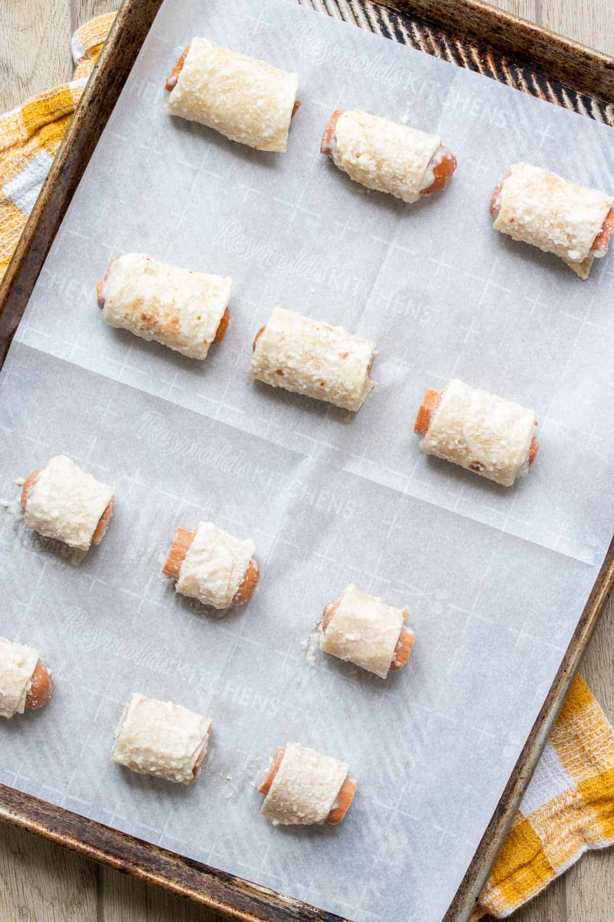 A baking sheet with parchment and hot dog pieces rolled with batter coated tortilla strips on it.