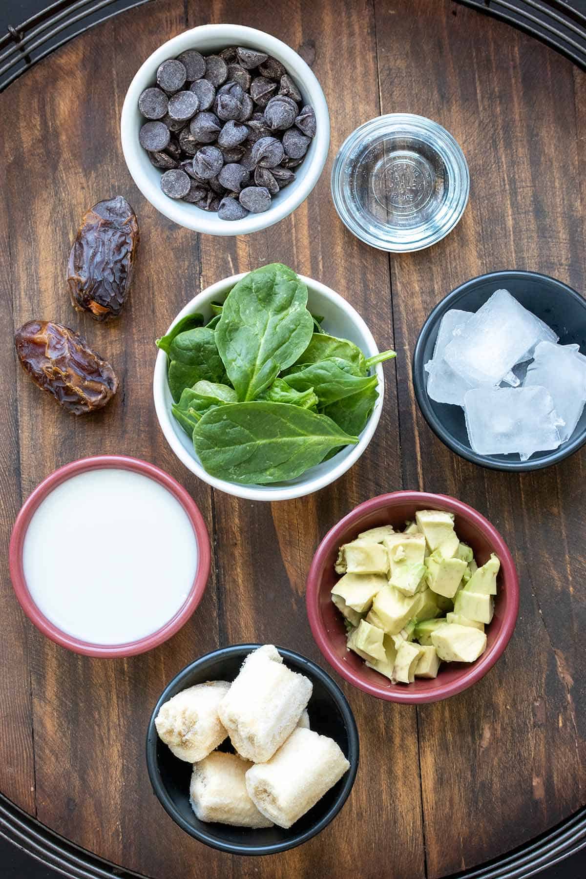 Different colors bowls with chocolate chips, clear liquid, ice, avocado, bananas, spinach and milk next to dates on a wooden surface.