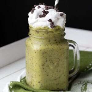 A green smoothie that is a shamrock shake in a glass mug topped with whipped cream and chocolate chips.