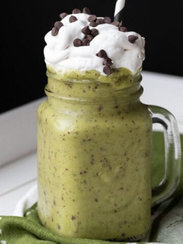A green smoothie that is a shamrock shake in a glass mug topped with whipped cream and chocolate chips.