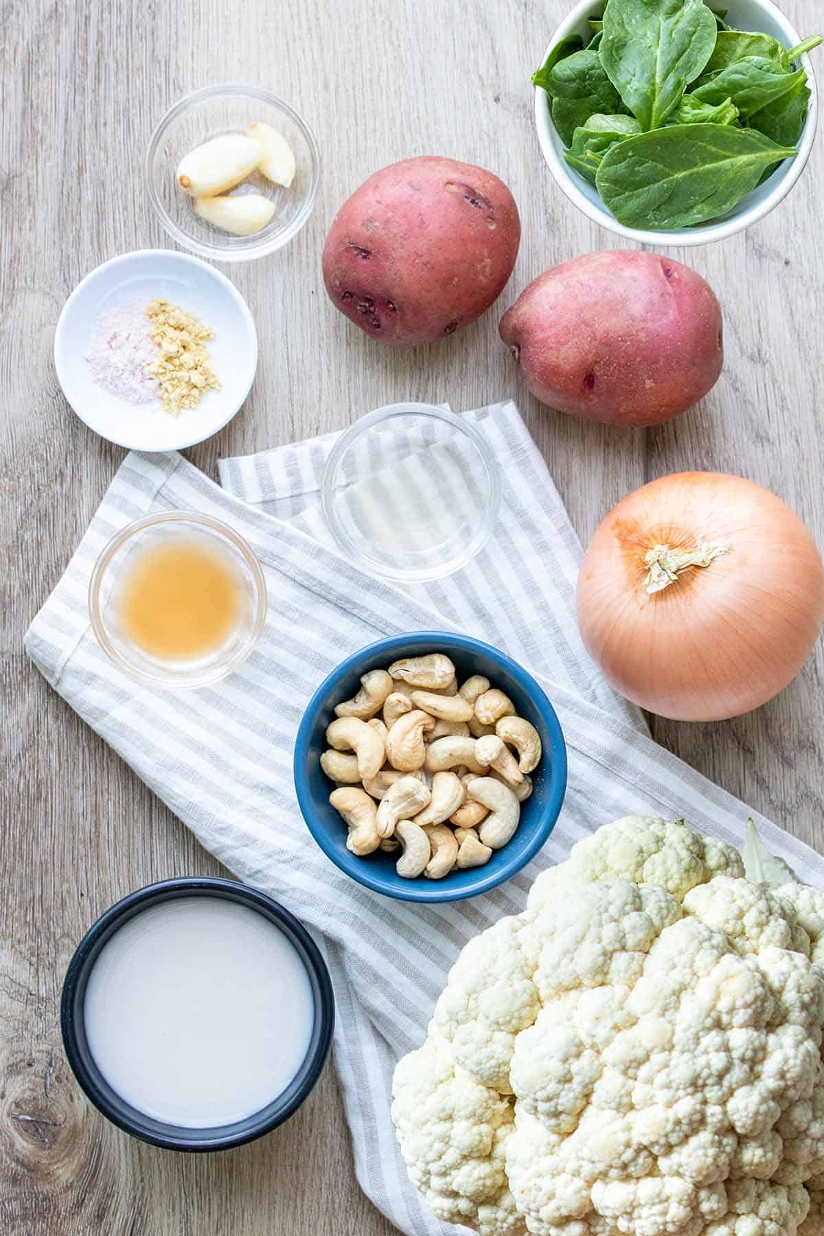 Bowls of milk, cashews, vinegar, lemon juice, spices, garlic and spinach on a light wood surface with cauliflower, onion and potatoes.