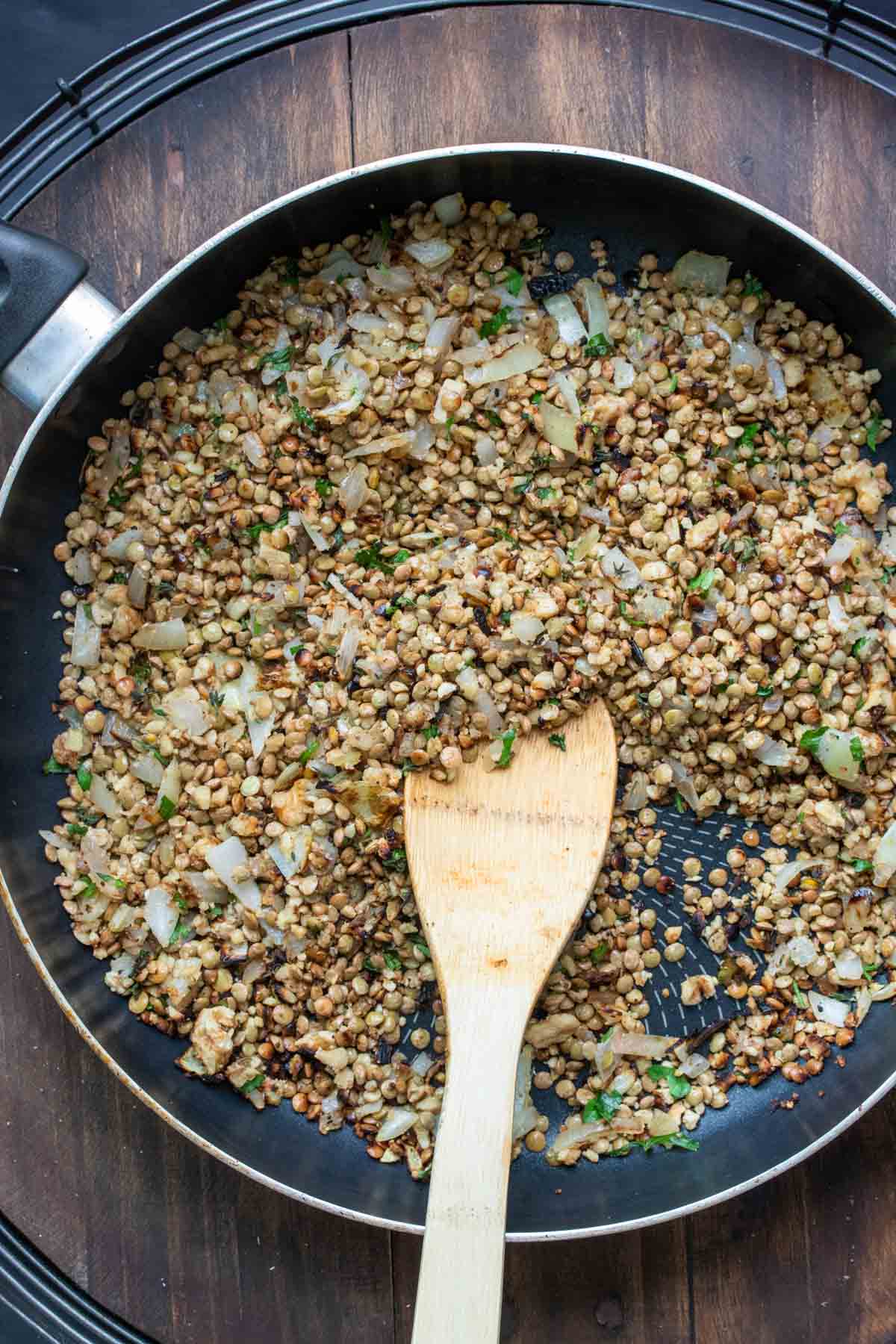 A wooden spoon sauteeing an onion, parsley and lentil mixture in a pan.
