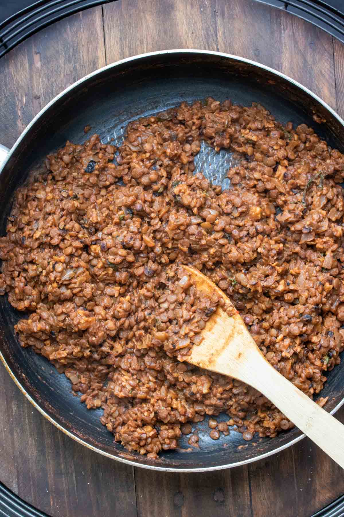 A wooden spoon mixing a red brown bean based crumble in a pan.