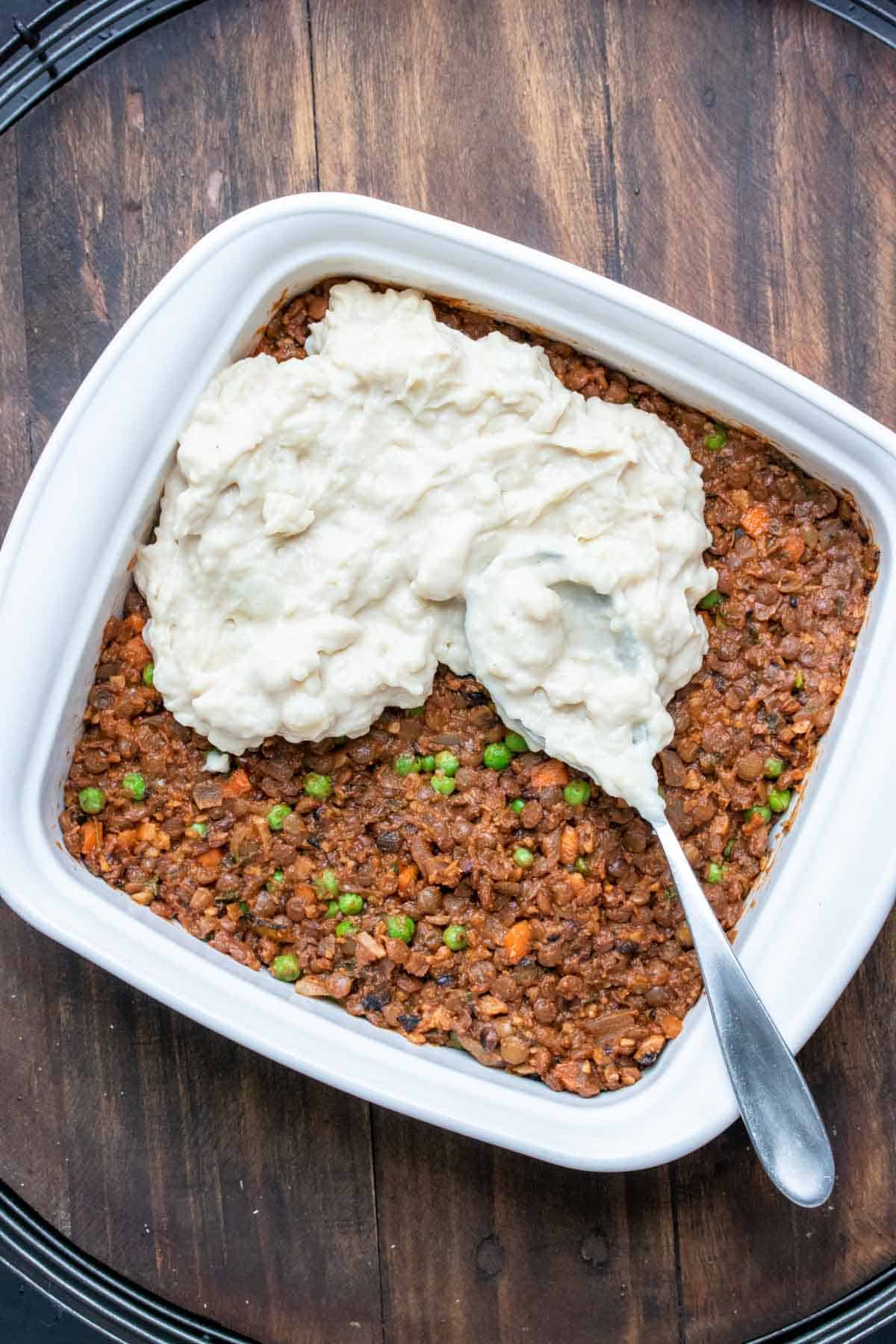 Top view of a white baking dish with a ground bean meaty filling with carrots and peas and mashed potato being spread on top.