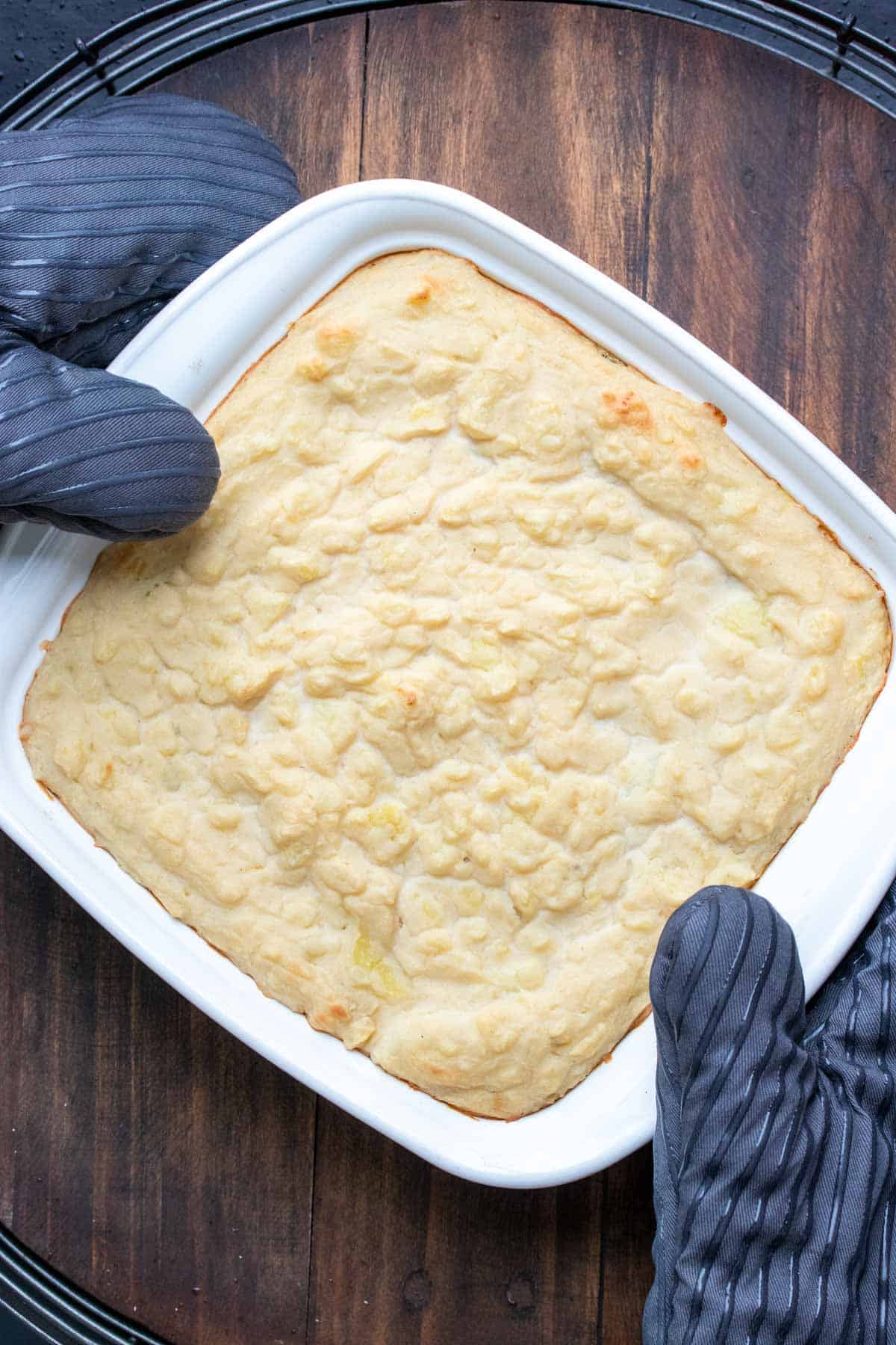 Two hands with oven mitts holding a white square baking dish with a baked casserole that has a potato topping.