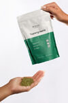 A green and white bag being held by a hand over another hand holding greens powder.