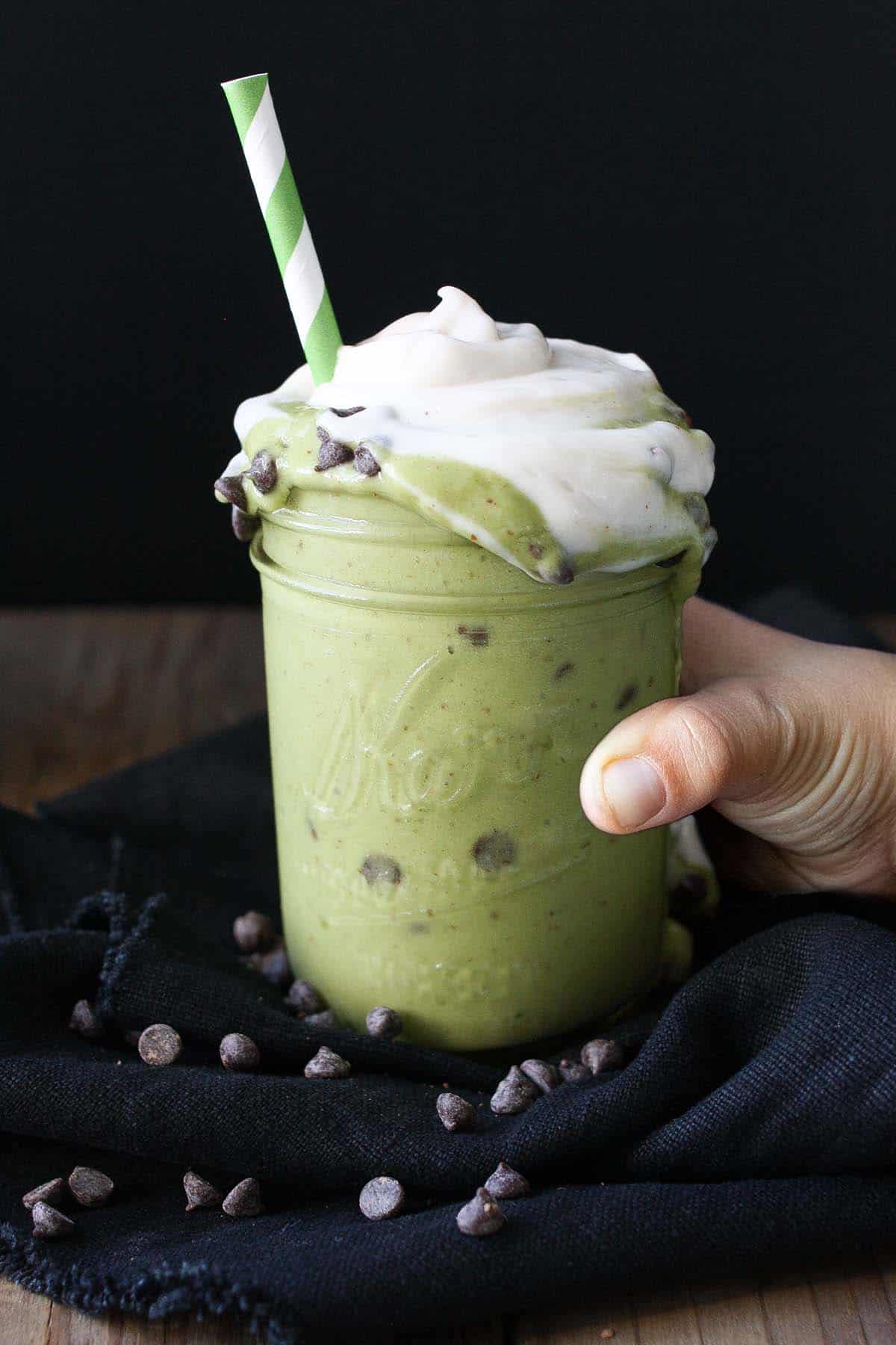 A hand grabbing a glass jar with a green shake in it with chocolate chips and whipped cream on top.