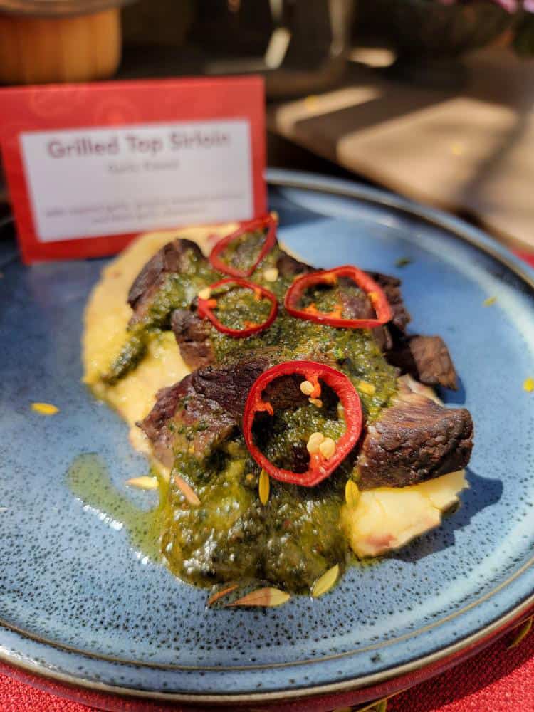 A blue plate with mashed potatoes topped with green sauce, red pepper slices and pieces of meat.