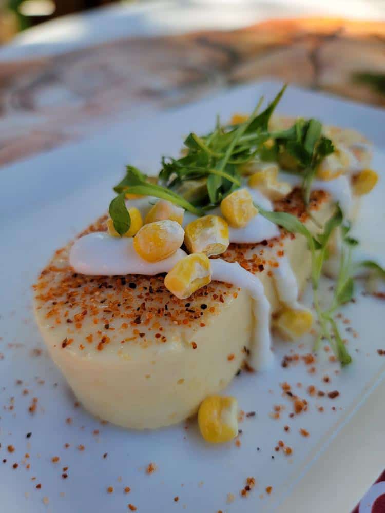 A yellow ice cream like bar with corn, cream and cilantro on top sitting on a white plate.