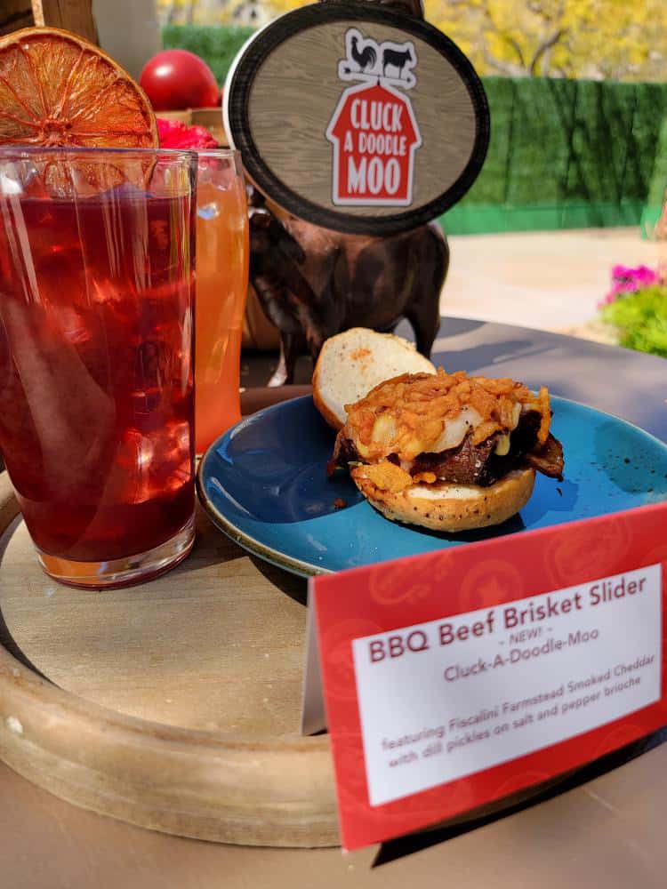 A blue plate with an open faced brisket slider on it topped with fried onions and drinks next to it.