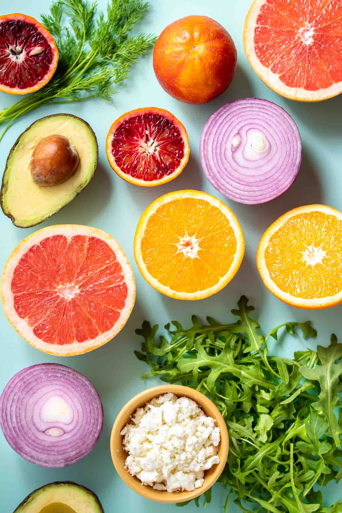 Turquoise background with grapefruit, orange, red onion and avocado halves on it and greens and a bowl of feta.
