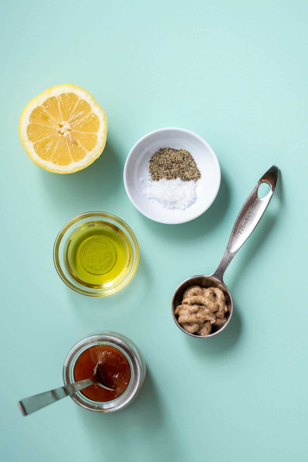 Half a lemon with bowls of oil, salt, pepper and honey and a spoon of mustard on a turquoise background.