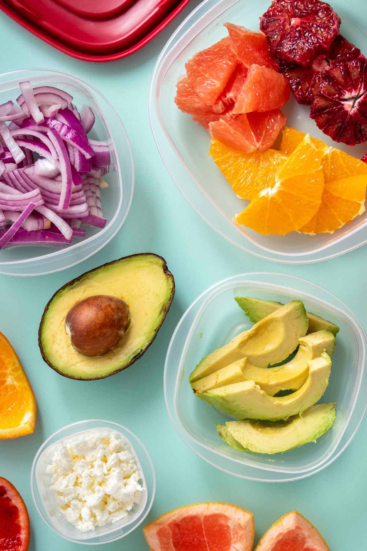 Plastic containers with citrus, avocado, onion and feta on a turquoise background.