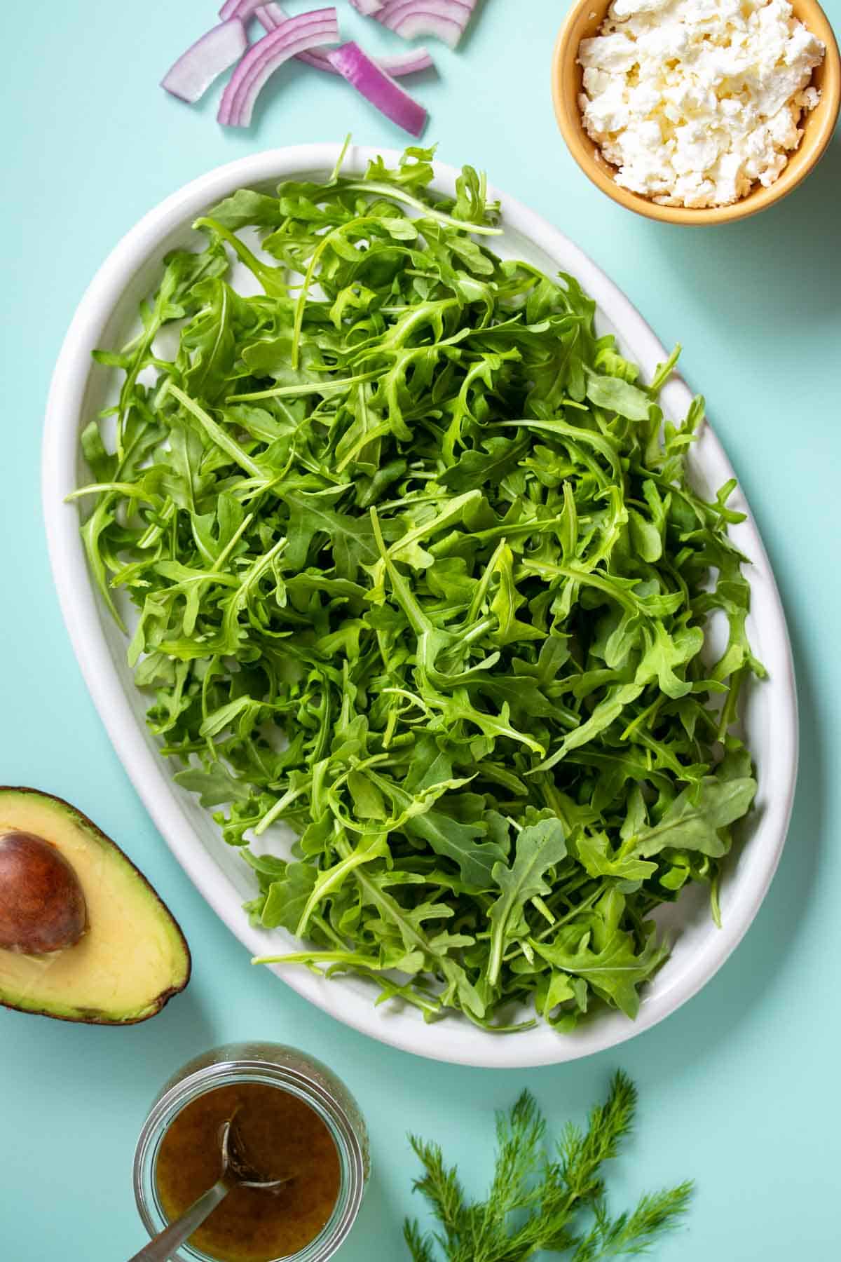 A white platter with arugula on it and other ingredients for a salad surrounding it on a turquoise background.